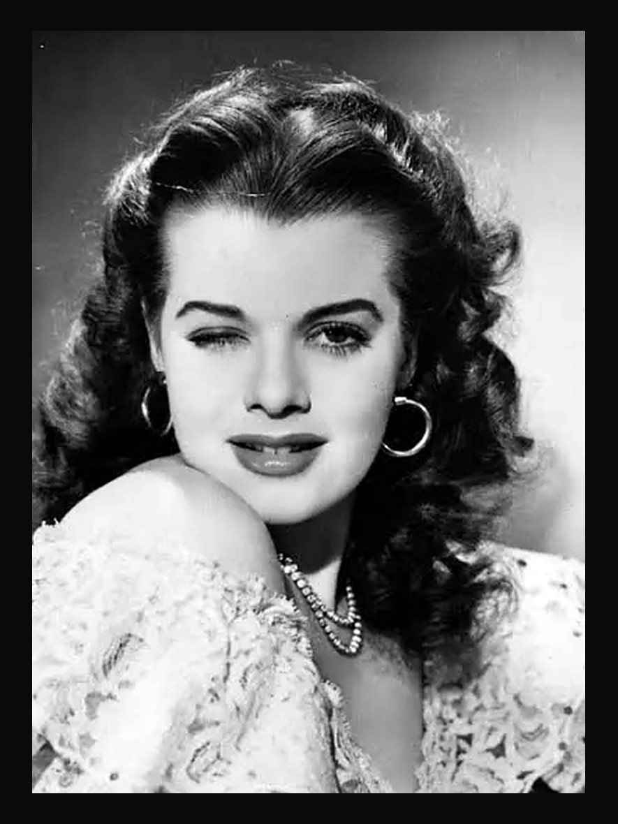 A studio publicity portrait of Janis Paige in a lace gown and hoop earrings
